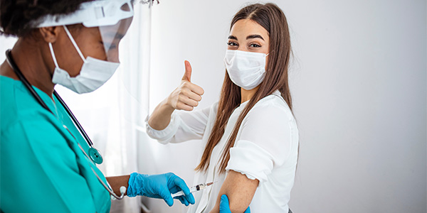 mulher recebendo dose de vacina e fazendo um sinal de positivo com a mão